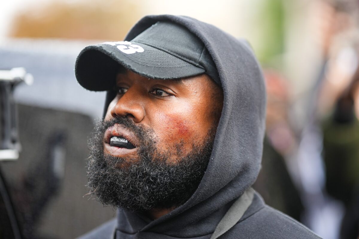 Un homme avec une barbe et une moustache porte un sweat à capuche noir sur un chapeau noir avec un protège-dents de boxe Balenciaga.