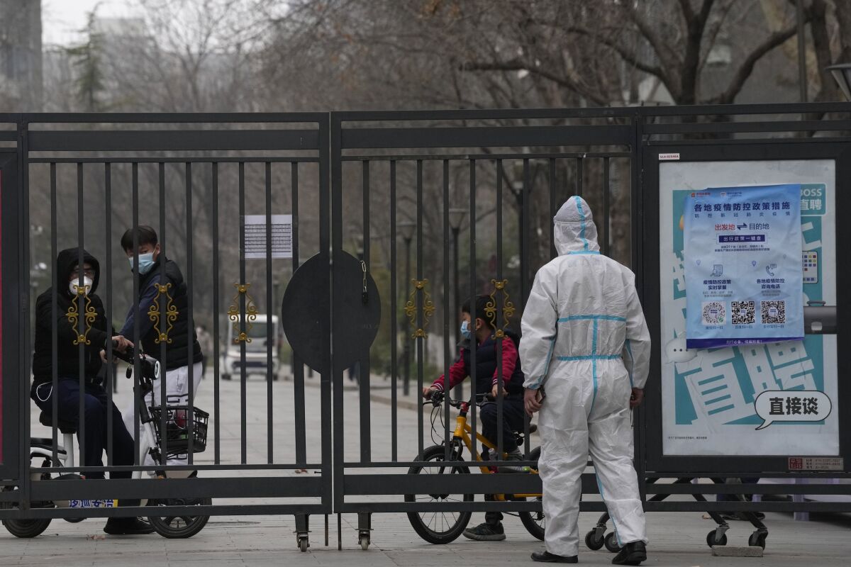 Residents in a locked down community look out from a closed gate as a worker in protective gear monitors access on Sunday, March 13, 2022, in Beijing. The number of new coronavirus cases in an outbreak in China's northeast tripled Sunday and authorities tightened control on access to Shanghai in the east, suspending bus service to the city of 24 million and requiring a virus test for anyone who wants to enter. (AP Photo/Ng Han Guan)