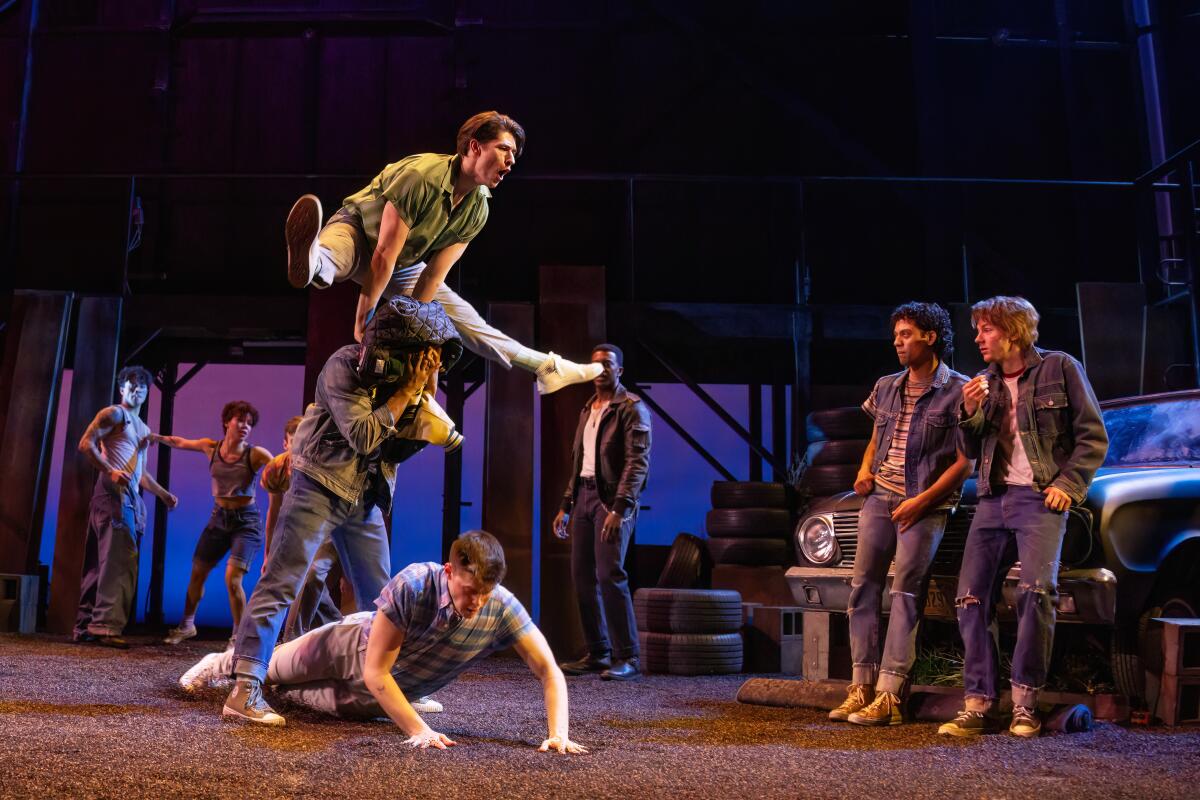 One person leapfrogs another as others stand watching and leaning against a car onstage