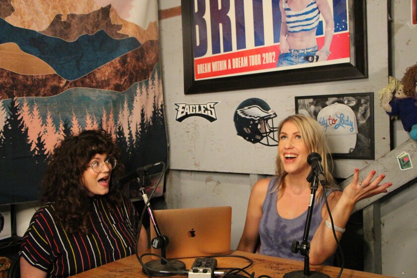 Babs Gray and Tess Barker sit at a table and speak into microphones. On a wall is a framed poster of Britney Spears.