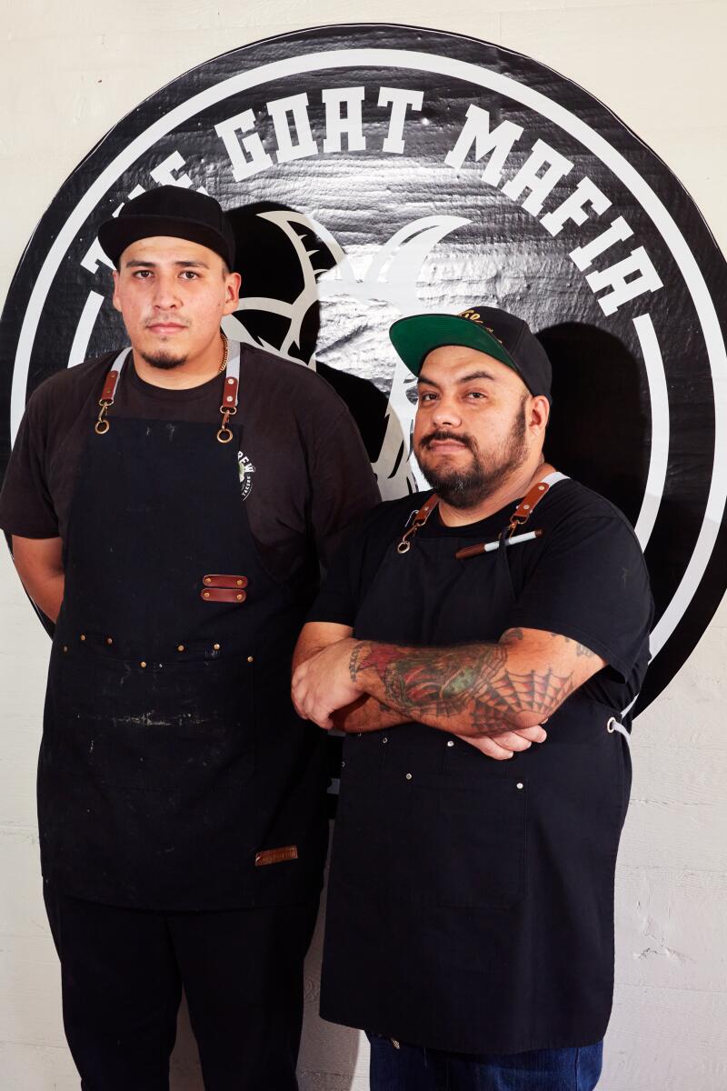 Chefs Iván Flores and Juan García, dressed all in black, stand in front of the Goat Mafia logo. 