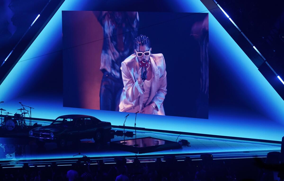 A man in white sunglasses in a suit holding a microphone and appearing on a large screen above a stage