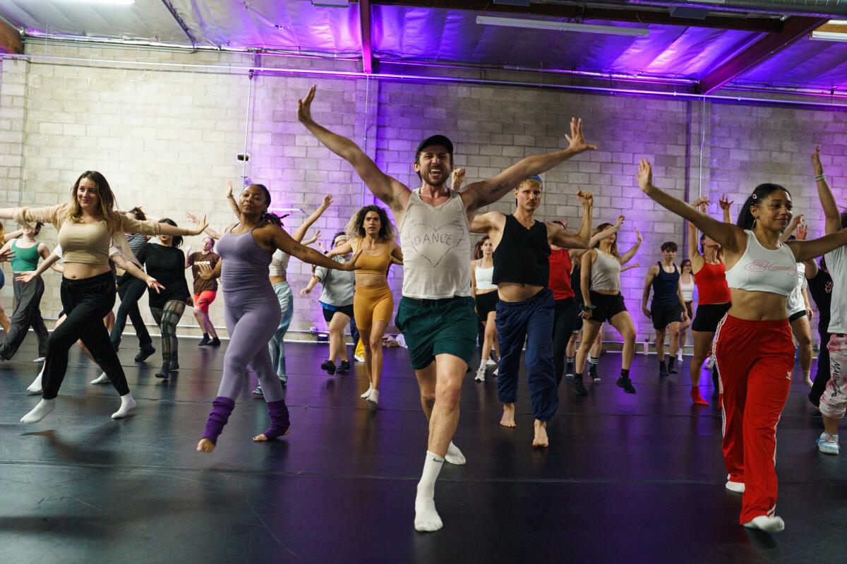 A man stands, arms raised, at the front of a class of people in workout clothes
