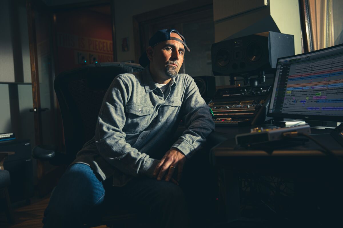 A man wearing a cap, button-down shirt and jeans leans on a recording studio control board.