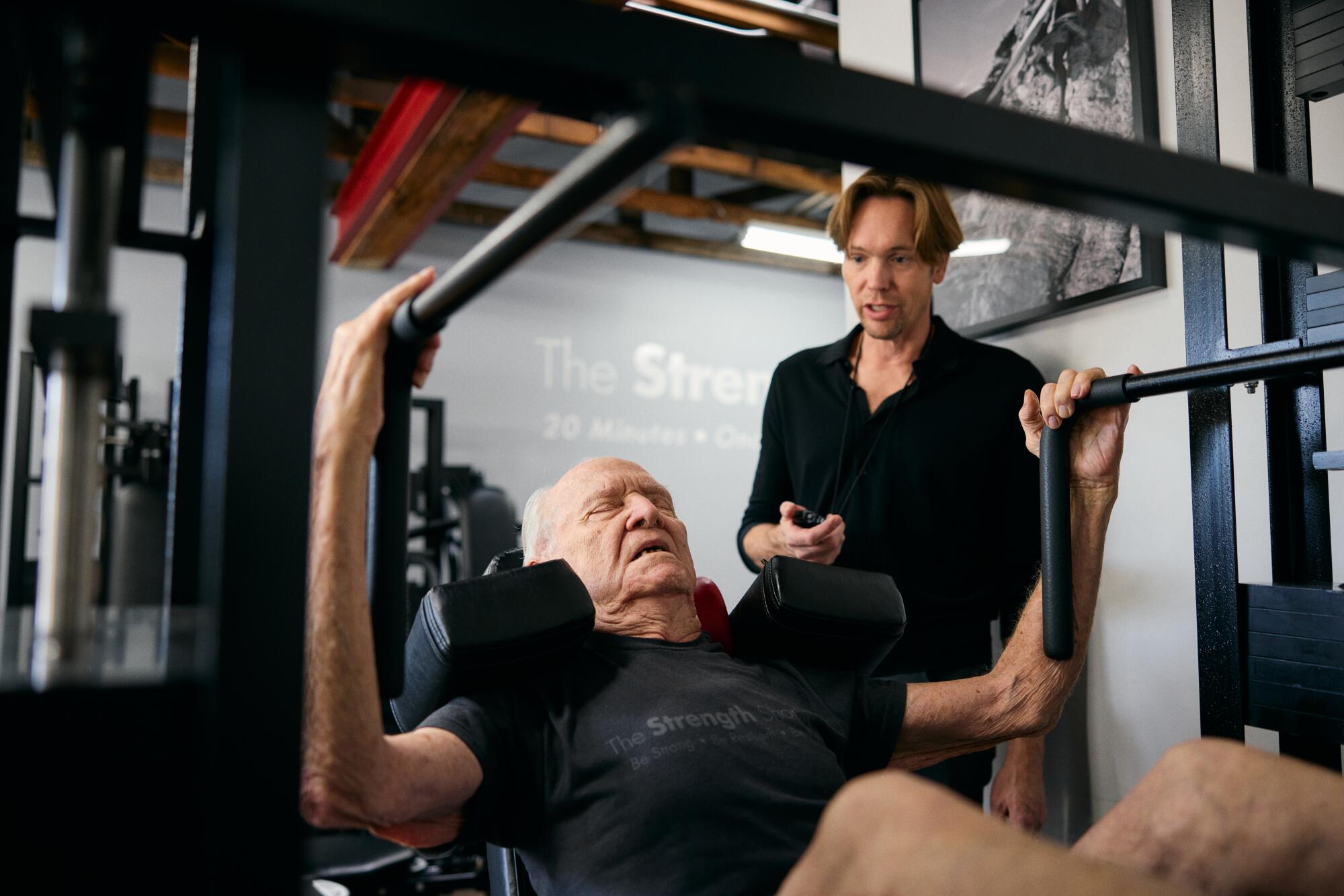 A 92-year-old man works out on the leg press machine as his trainer watches.