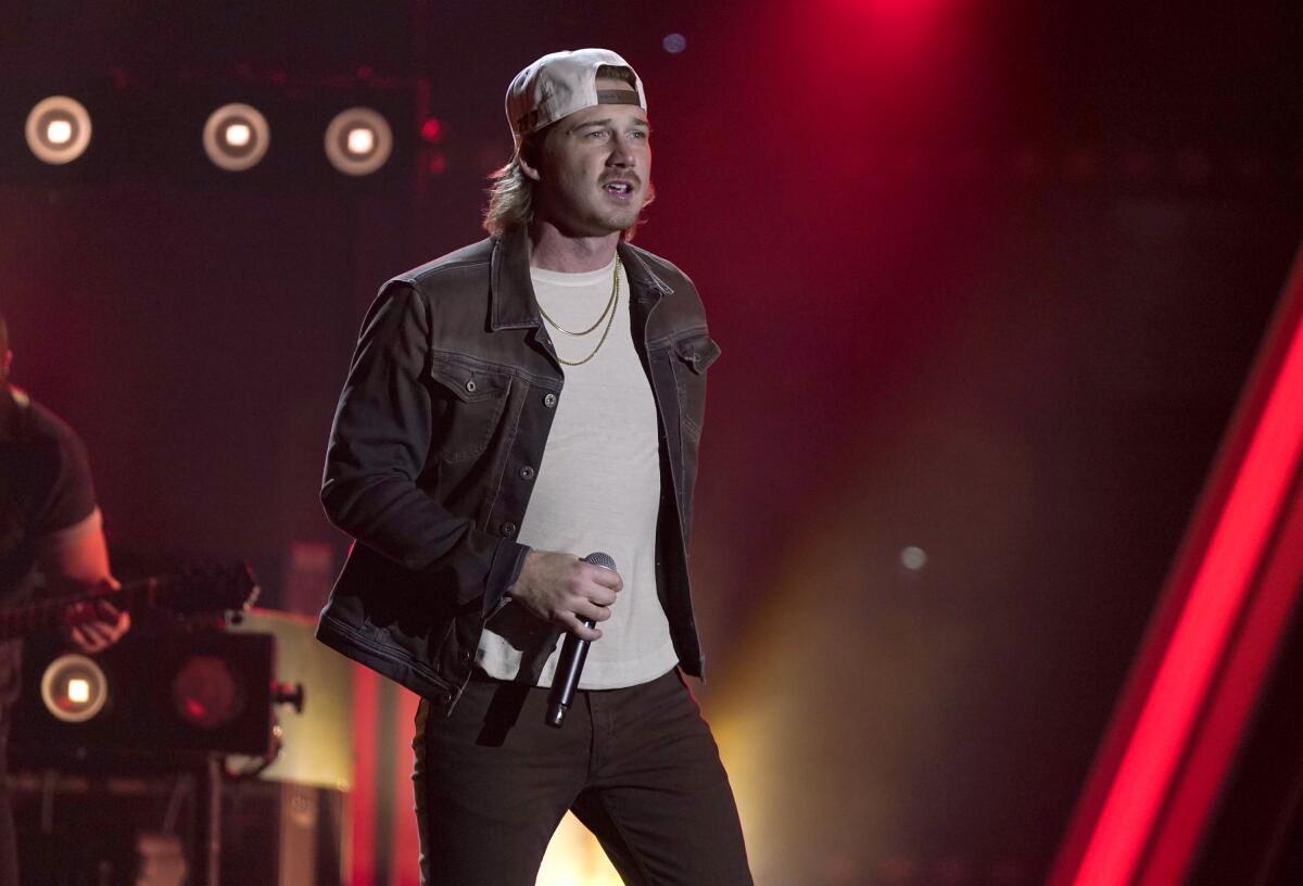 A man performs onstage, holding a mic and wearing a backwards white hat, black jacket, white T-shirt and dark pants. 
