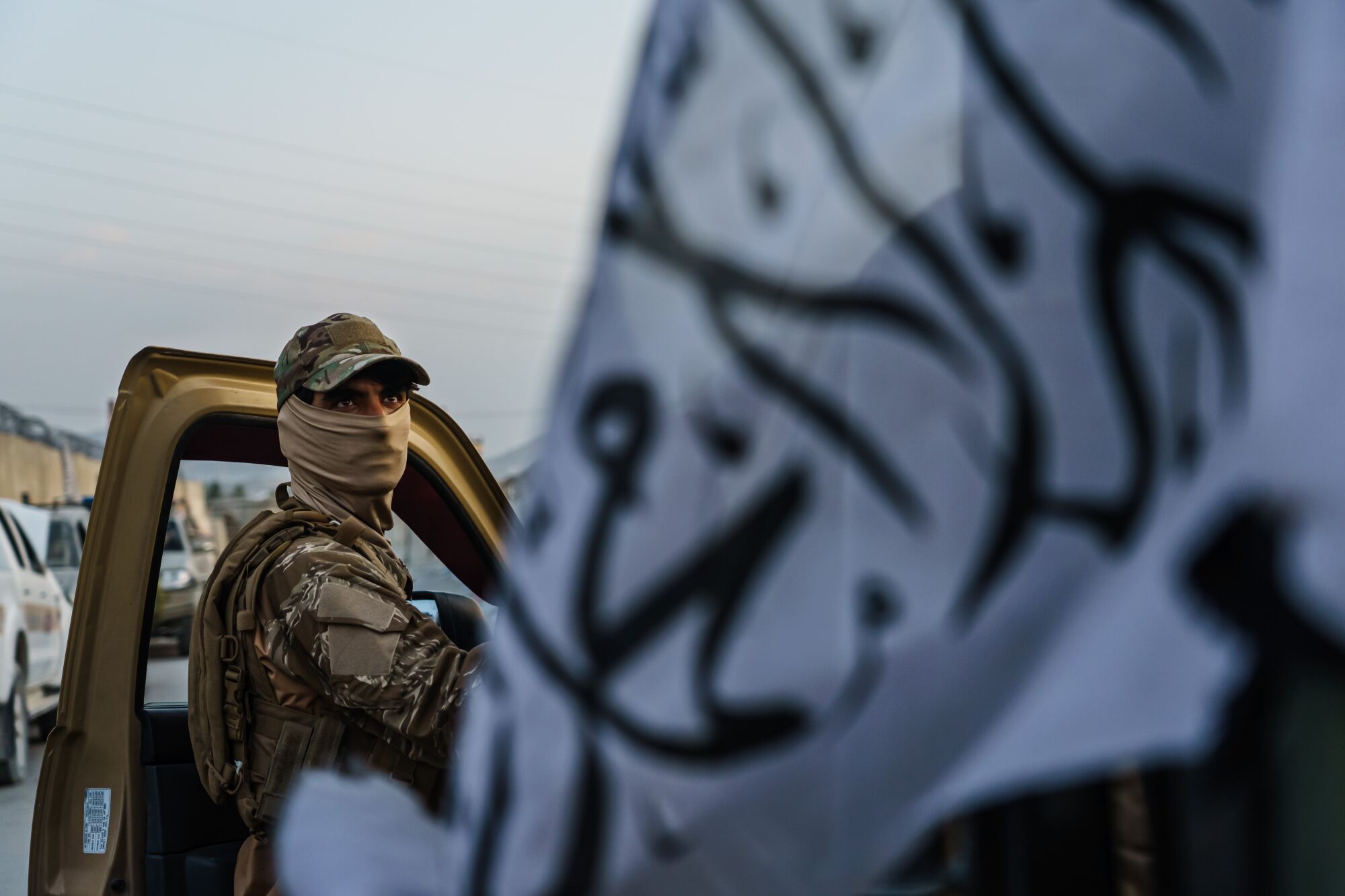 A Taliban fighter appears in the open door of a vehicle next to a white Taliban flag