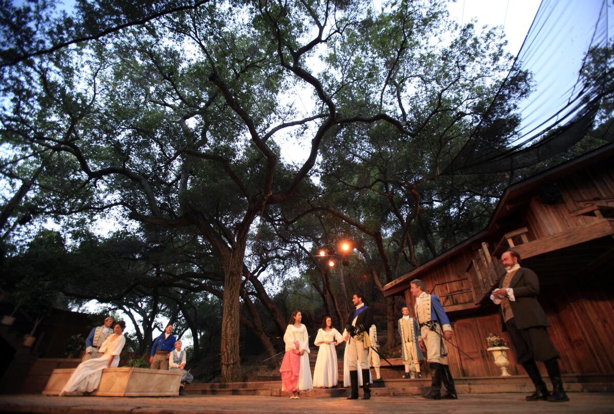 Actors in period dress stand on an outdoor stage underneath a canopy of trees