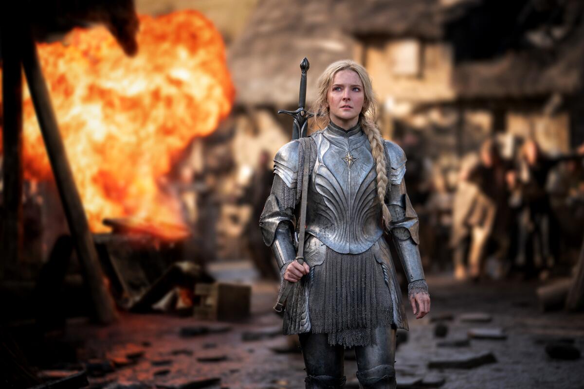A blond woman wearing armor walks toward the viewer with a sword over her shoulder as fire erupts behind her