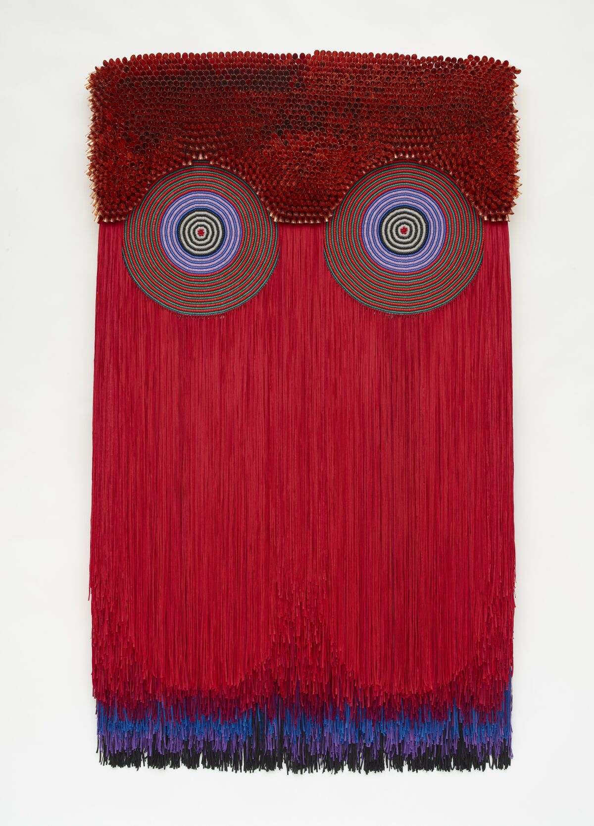 A vertical wallhanging features a row of glass beads and two circles over a curtain of red fringe