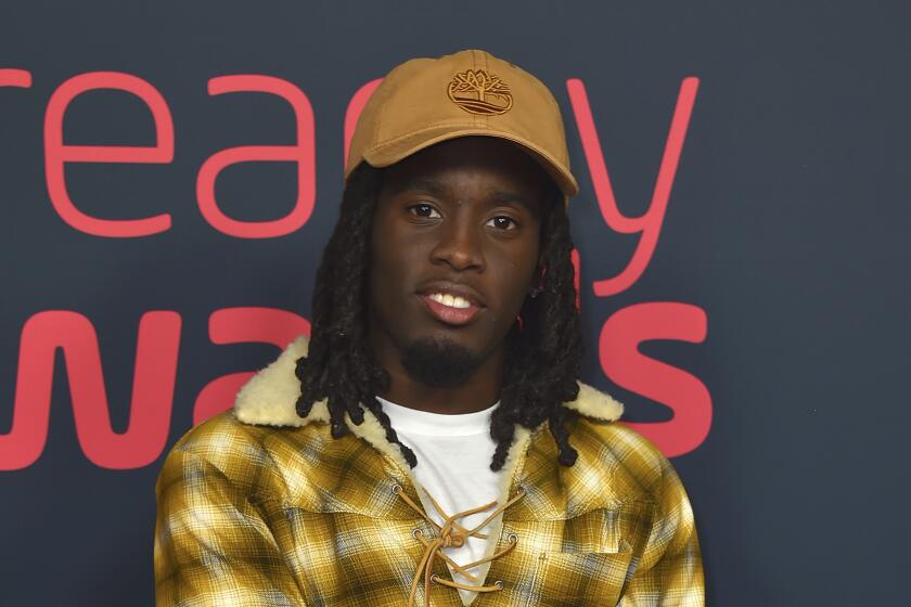 A young Black man with braids in a brown canvas hat and a yellow plaid jacket posing at a red carpet