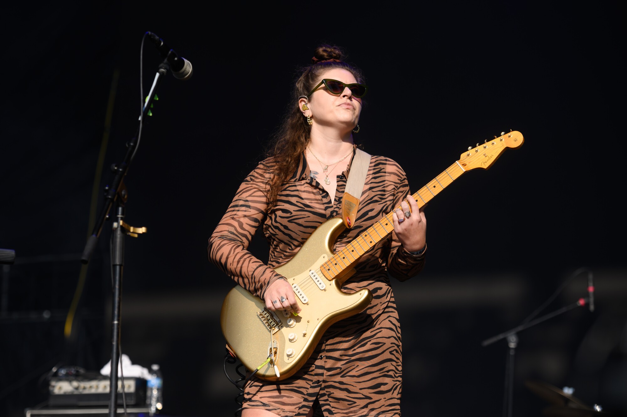 A woman onstage playing electric guitar, dressed in a leopard-print jumpsuit and wearing sunglasses.