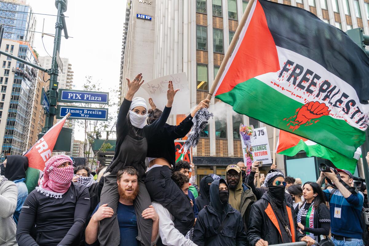 A man carries a woman on his shoulders amid a crowd on a city street as others wave "Free Palestine" flags or wear keffiyehs