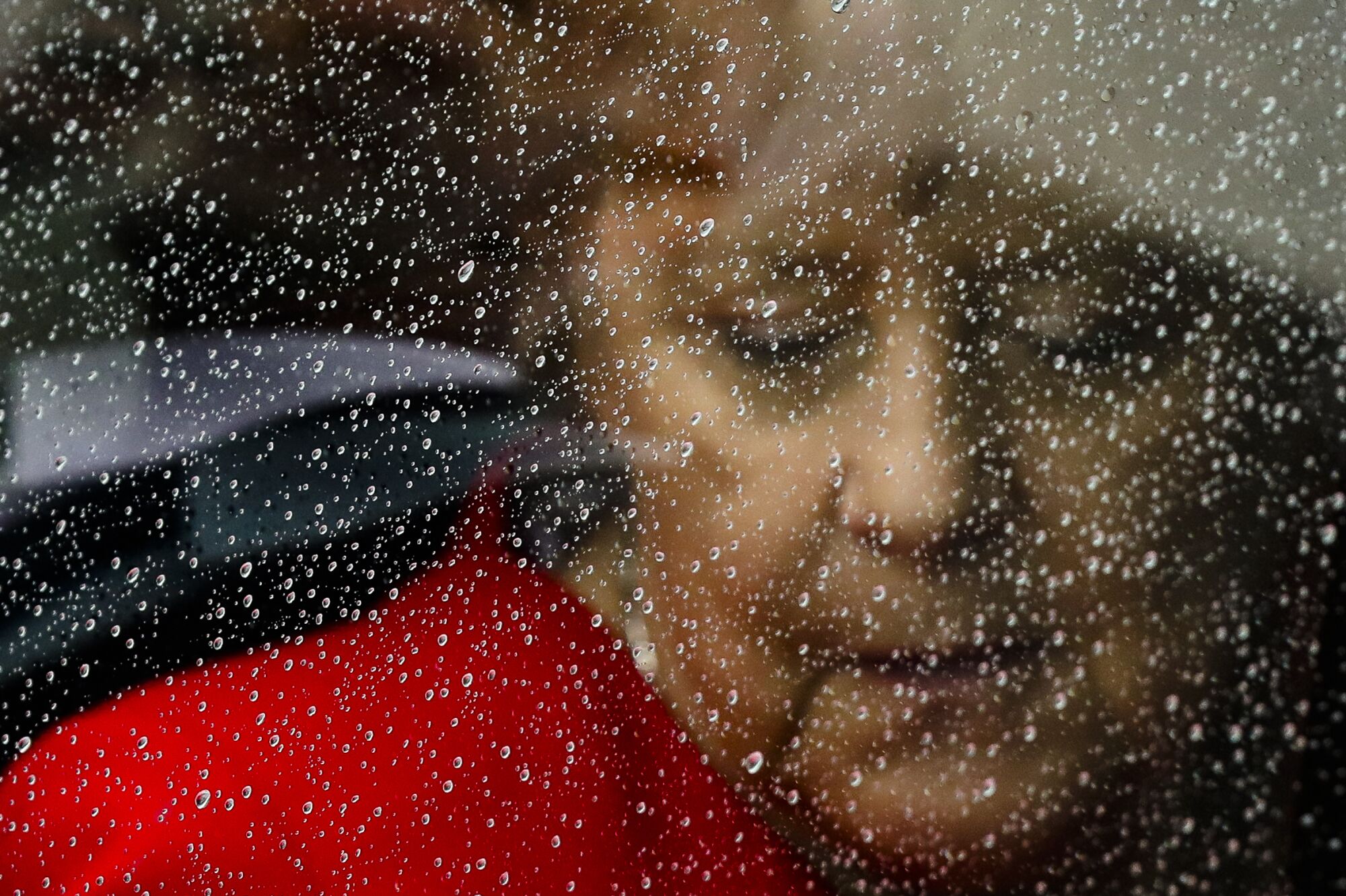 Angela Merkel, in a red jacket, looks down behind a rain-covered car window 