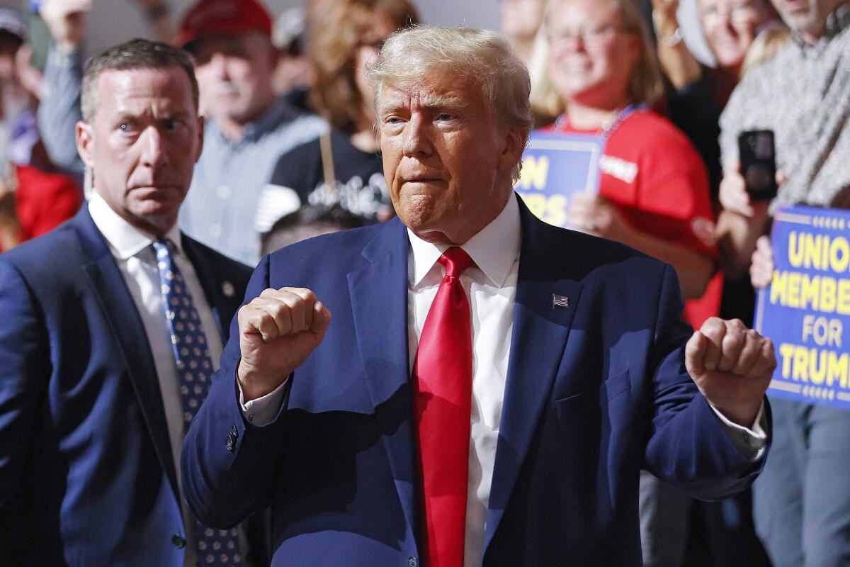 A man with blond hair, in blue suit and red tie, gestures with fisted hands near people holding campaign signs