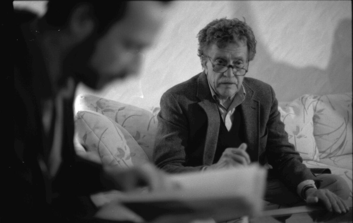 A black-and-white photo of a man in a sweater and blazer and glasses on a couch, write, and a blurry man, left 