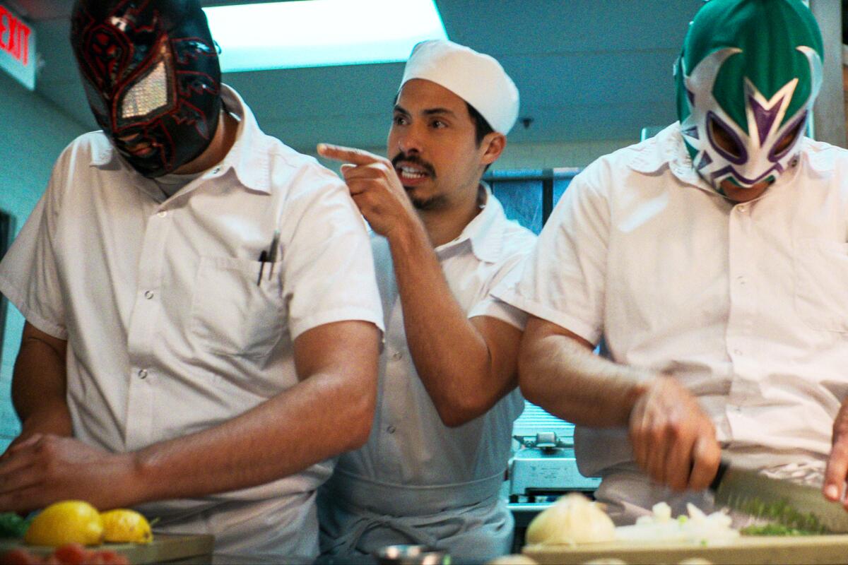 The character of Chris Morales stands between two cooks decked out in Mexican wrestler masks