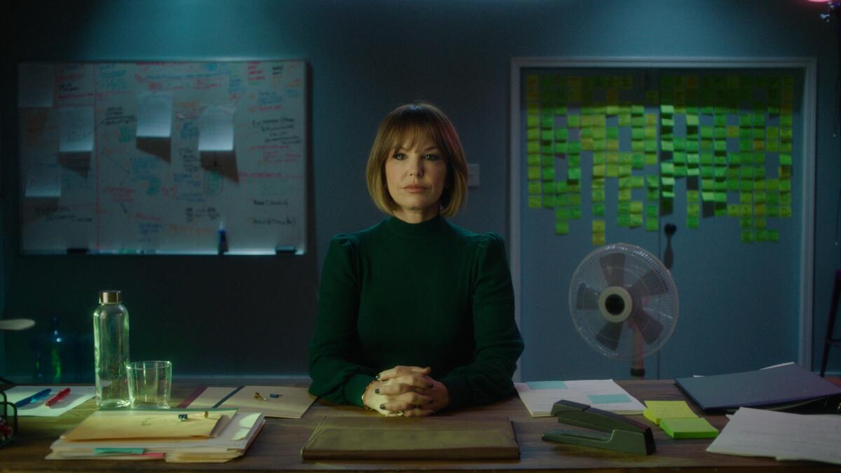 A woman in a green turtleneck sits at a desk with her hands clasped in front on her.