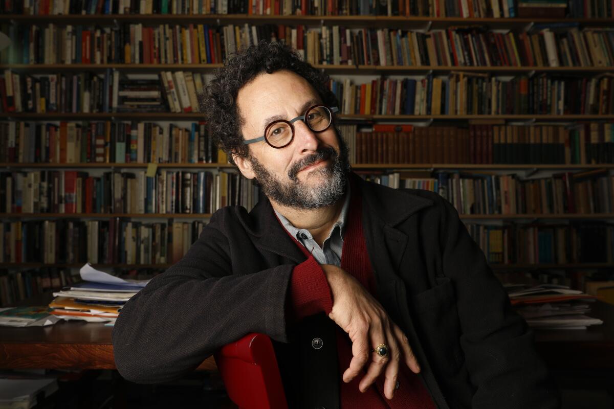 A man with round glasses and a short beard with a wall of books behind him.