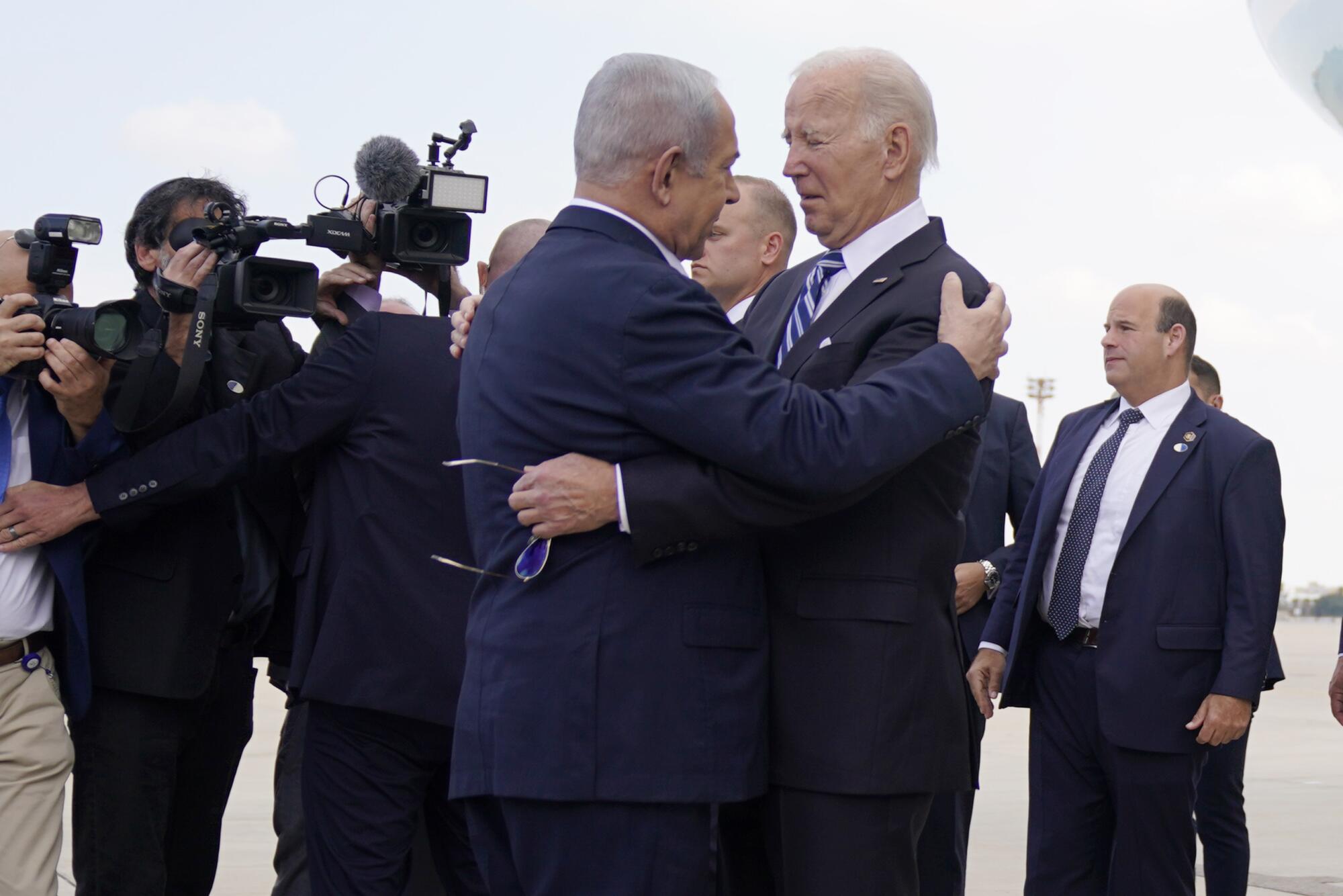Two men in dark suits embrace near photographers 