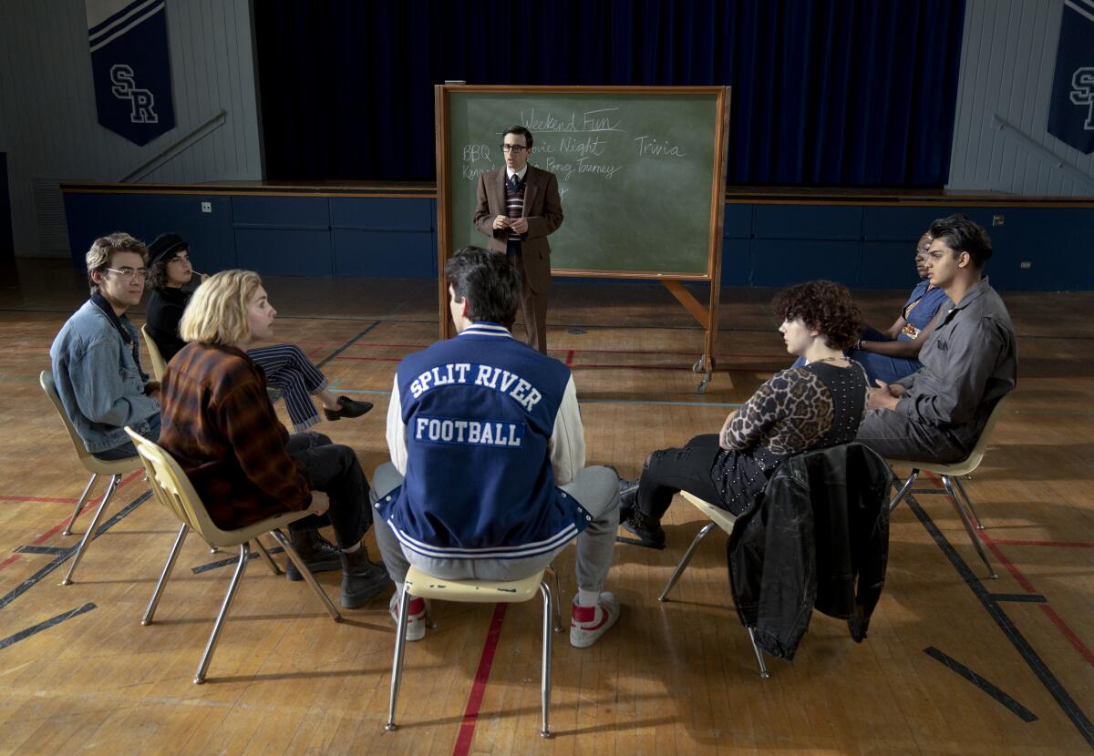A teacher stands at a blackboard in a school gym, facing a semicircle of students.