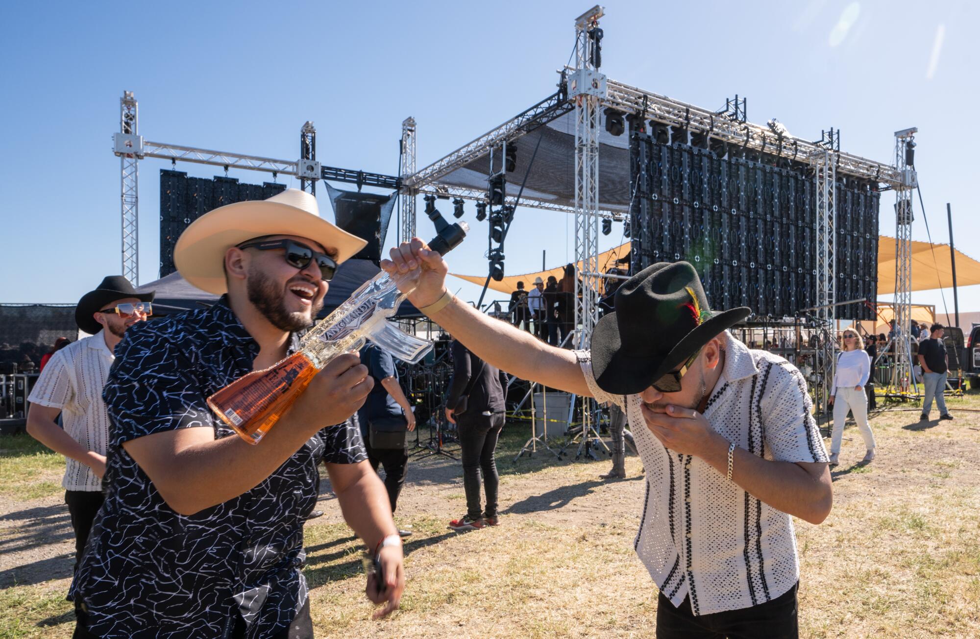 A man holds an AK-47-shaped tequila bottle as another man holds the end and puts his hand over his mouth after a swig. 