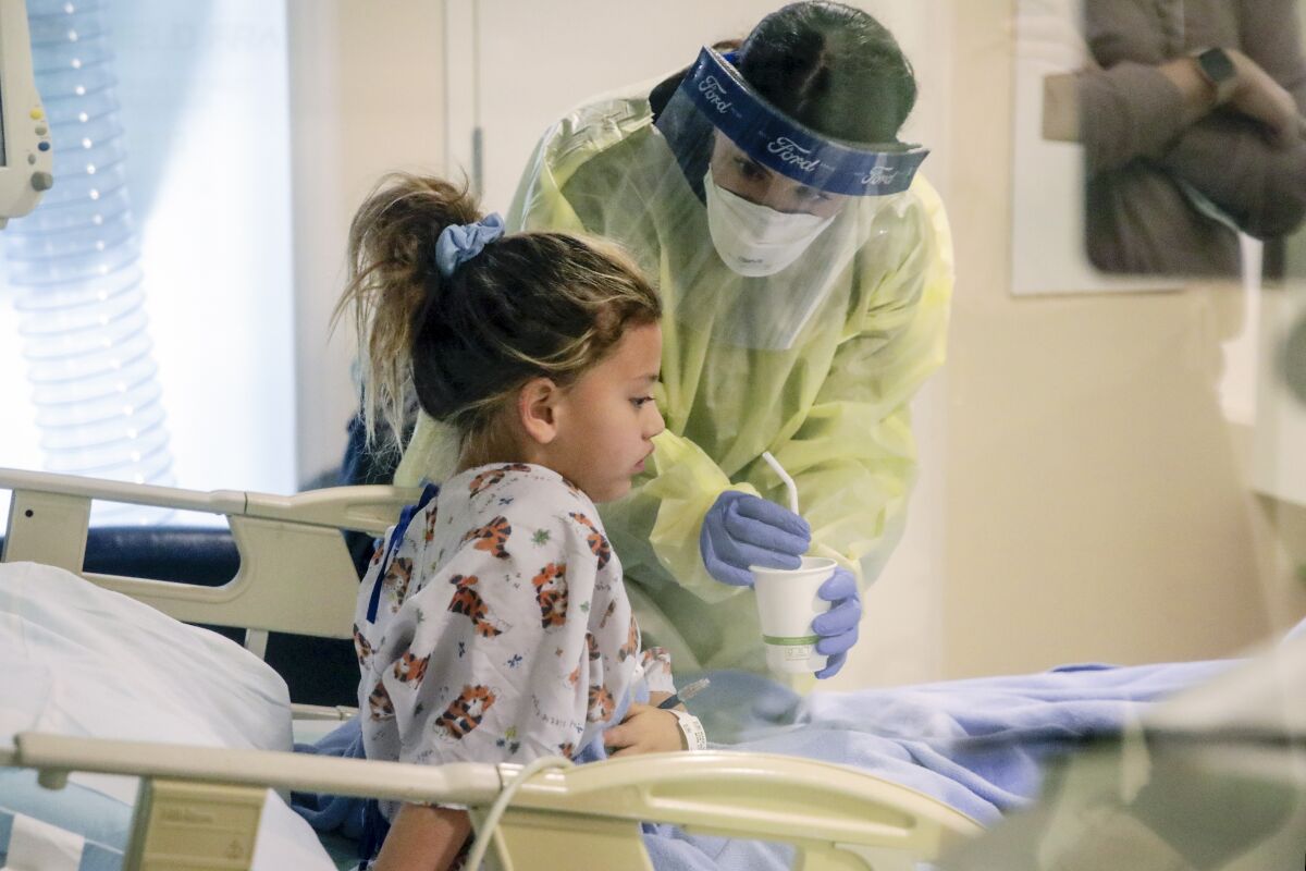 A person in mask and other protective gear offers a cup with a straw to a child sitting in a hospital bed.