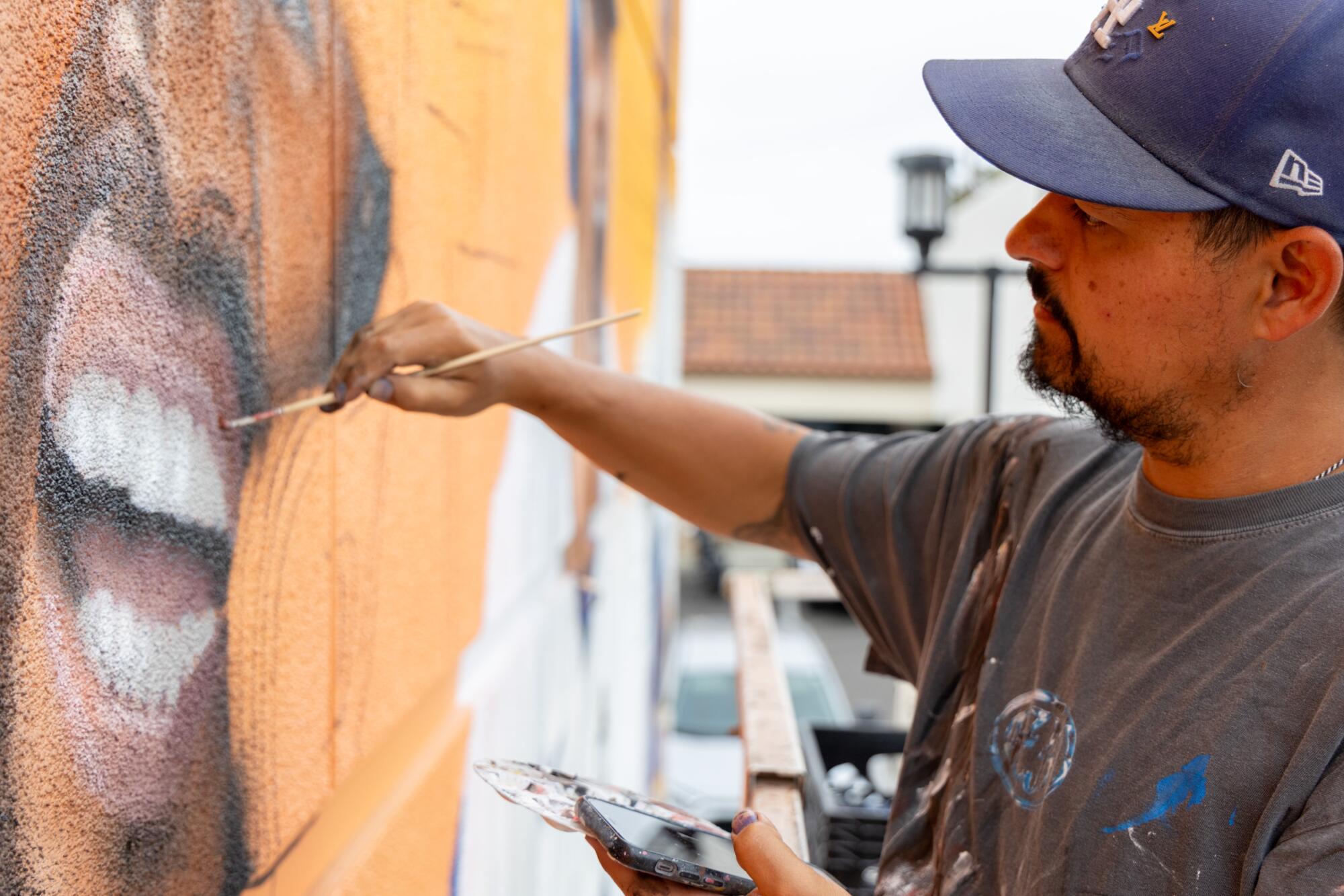 Um homem no boné de beisebol e uma camiseta pálida colocou uma paleta em uma mão e coloca um pincel no outro quando o mural é pintado