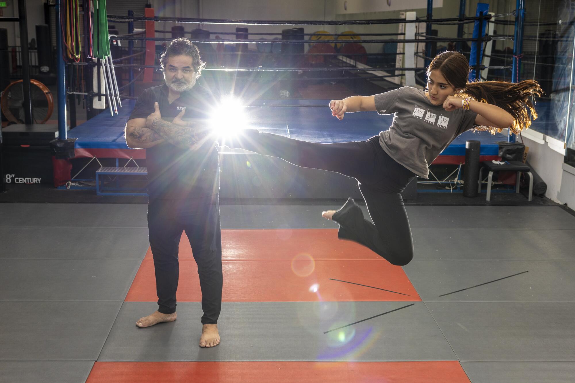 A woman does a jumping kick in the air next to a man in a dojo as sunlight enters the building.