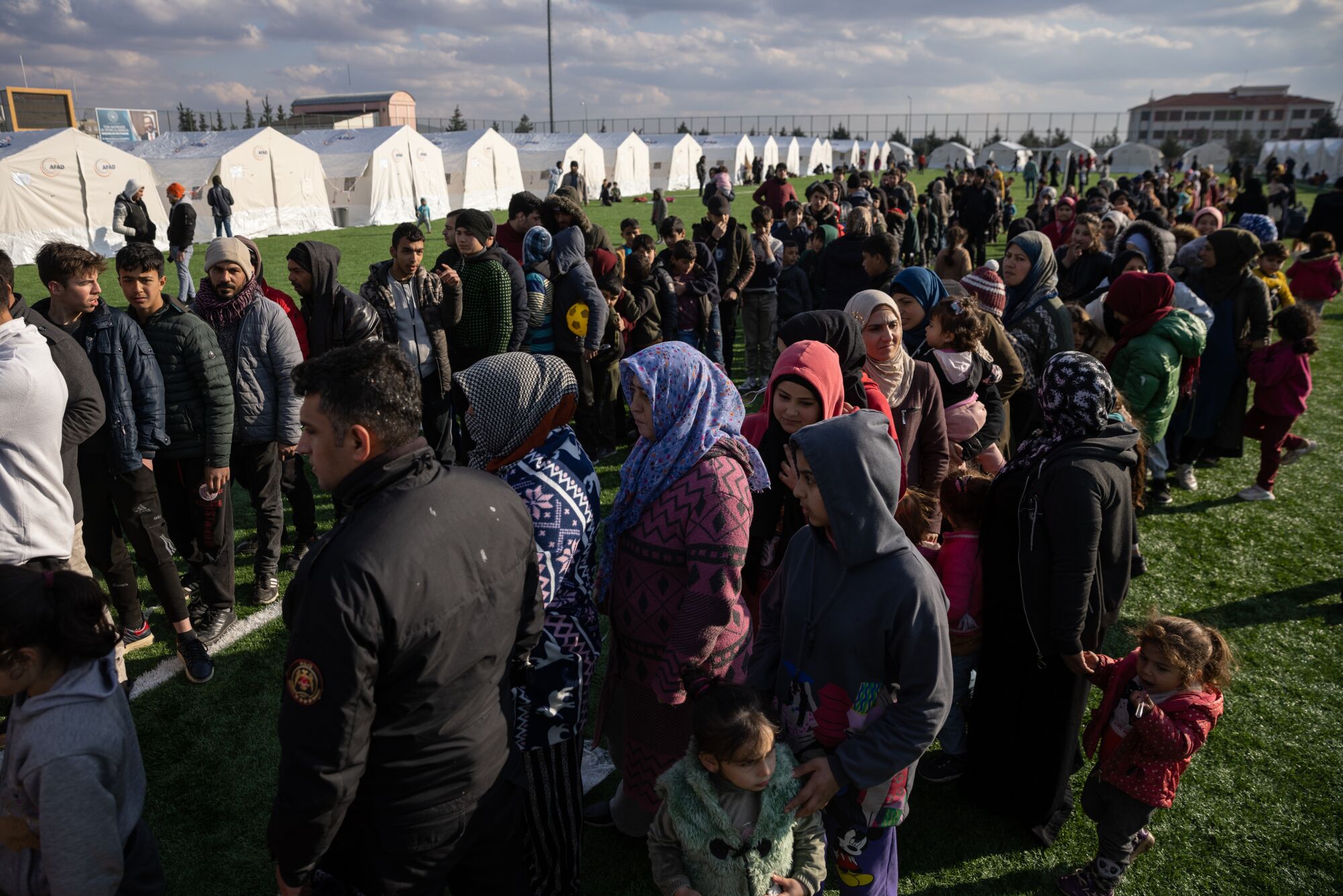 Displaced Syrians stand in line for food next to a row of tents.
