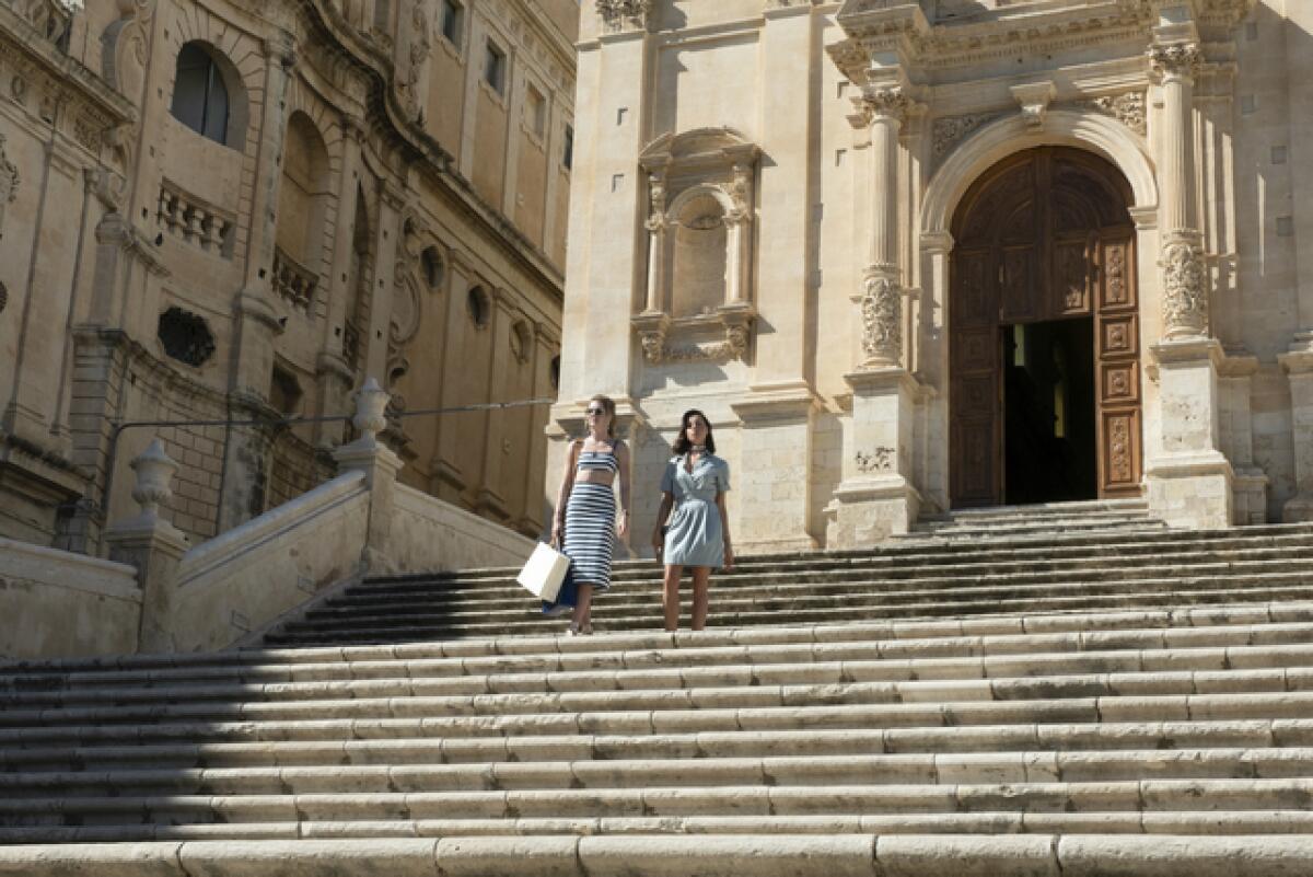 Two women with shopping bags stand on the steps of a church in "The White Lotus."