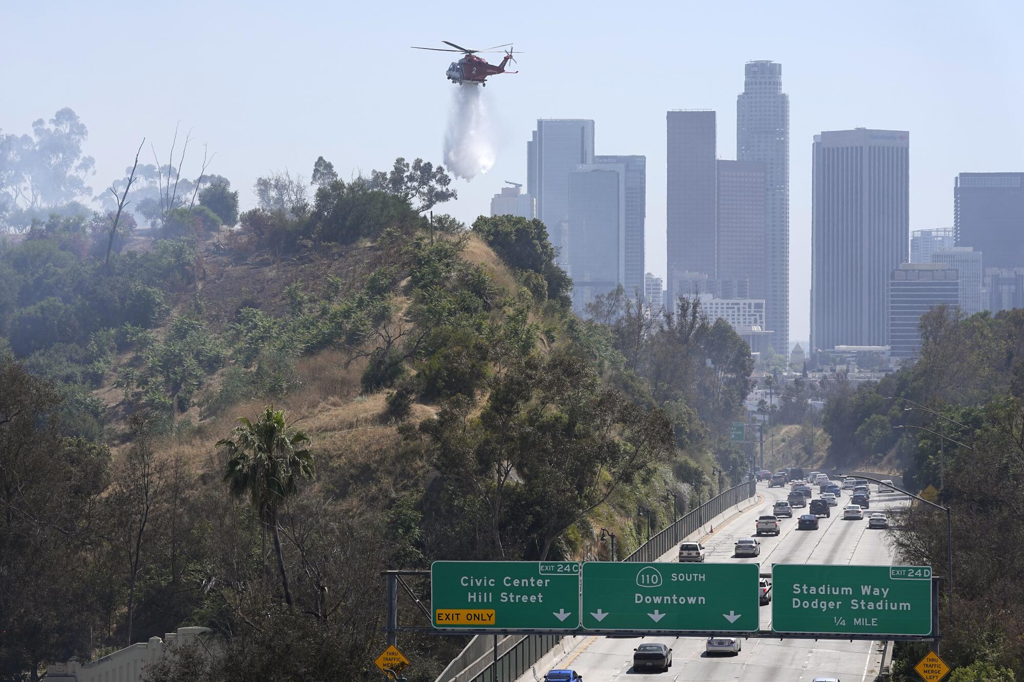 A helicopter drops water on a hillside against a backdrop of skyscrapers, near freeway signs for the 110 South to Downtown.