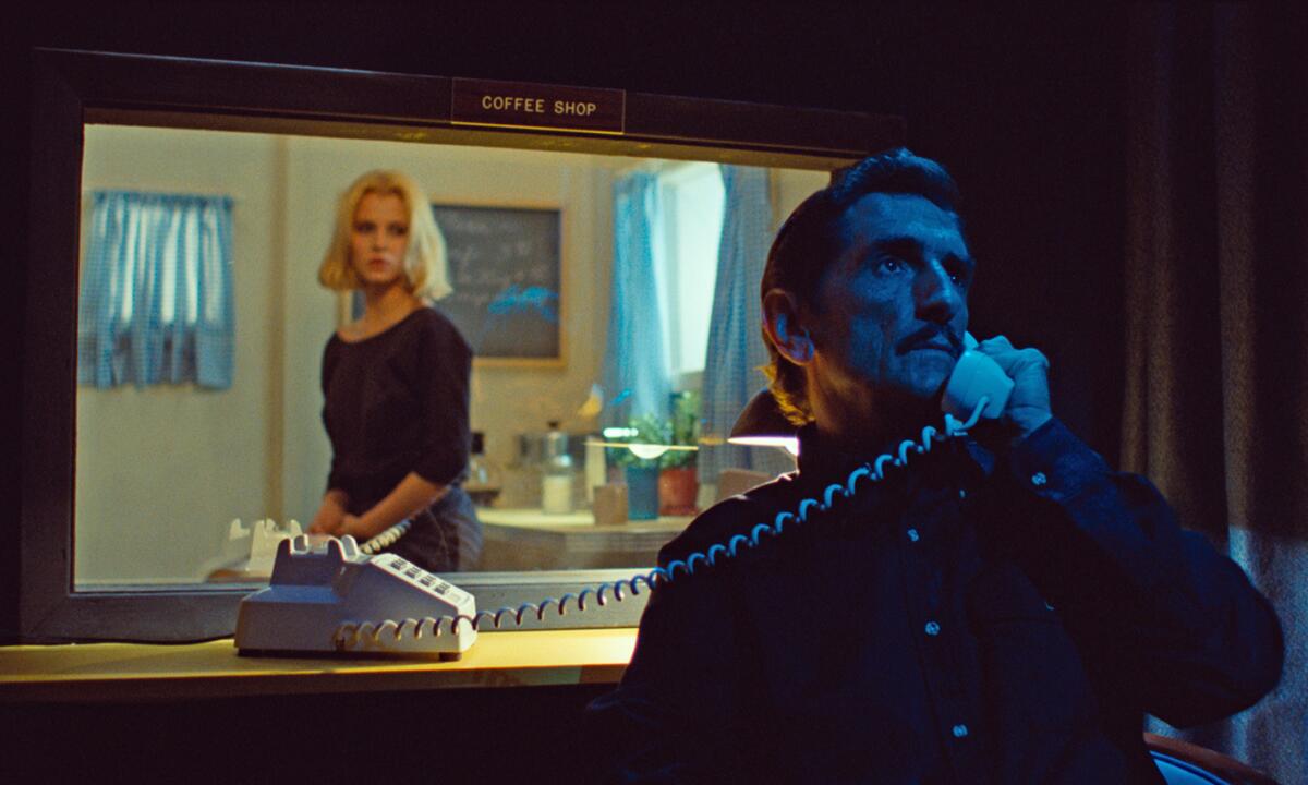 A woman and a man speak on a phone, separated by glass.