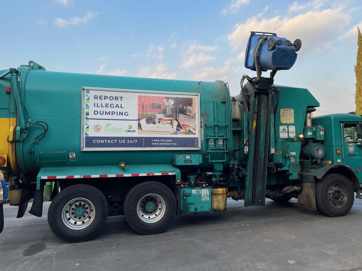 A green Los Angeles sanitation truck empties a blue recycling bin on a residential street