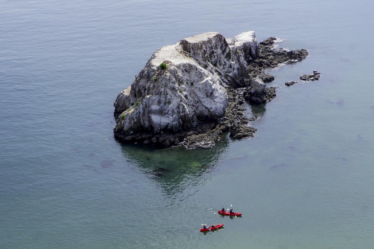 People in small boats in the ocean around a large rock