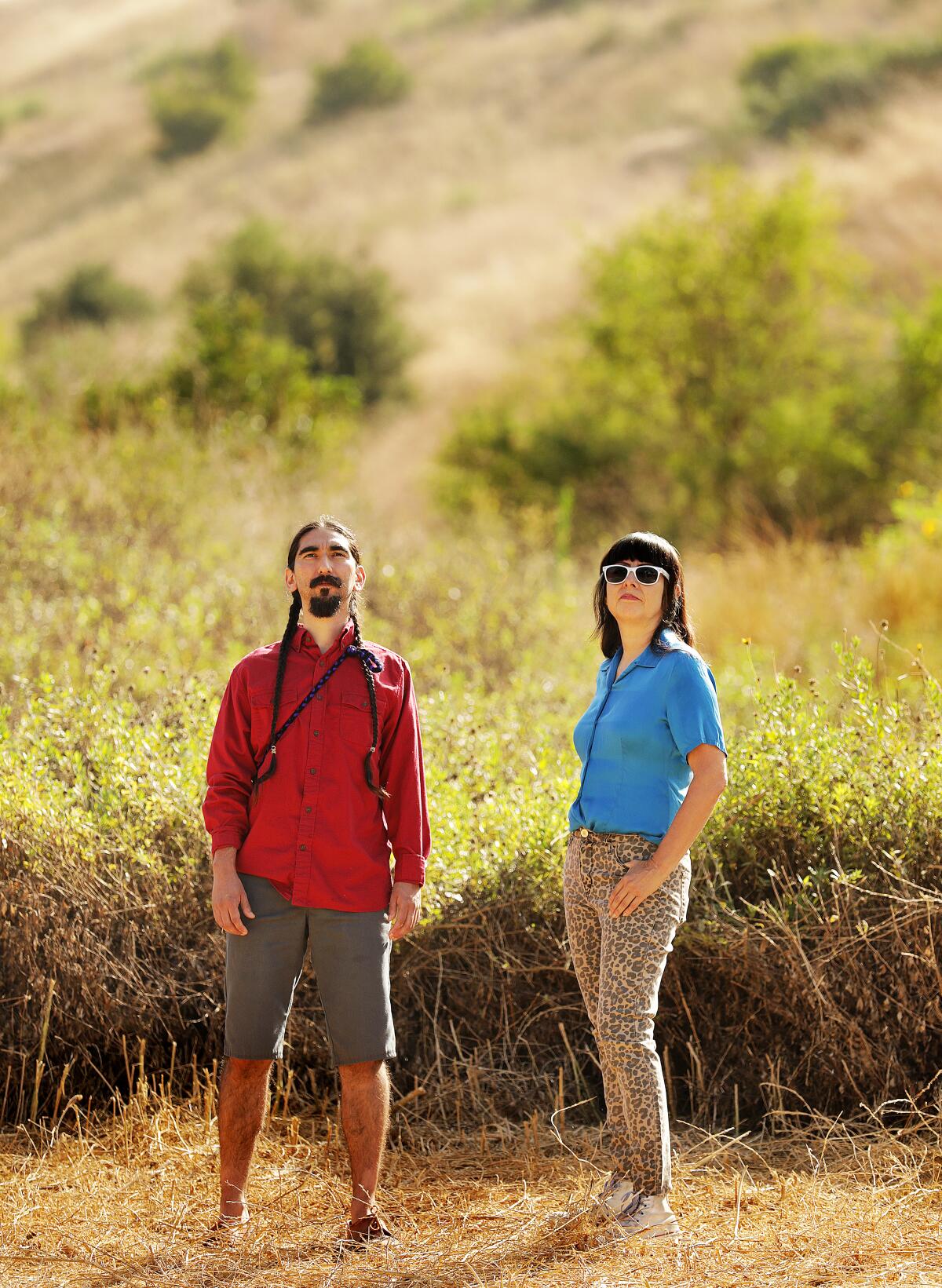 Artists Arturo Ernesto Romo and Sandra de la Loza stand before a grass-covered hillside at L.A.'s Ascot Hills Park