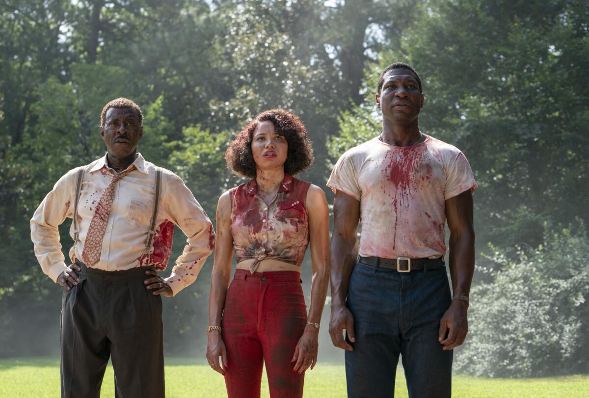 Three people in blood-spattered clothing stand in a grove of trees