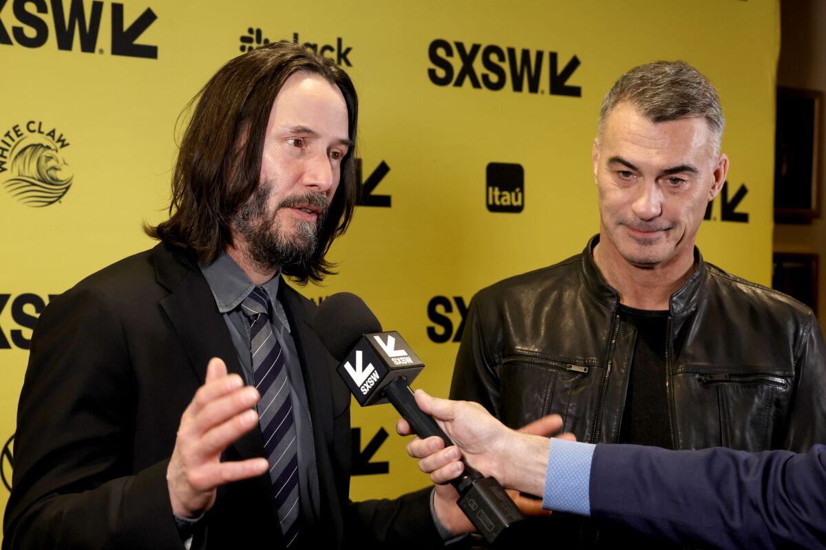 Keanu Reeves, left, and Chad Stahelski near a yellow backdrop with various brand logos