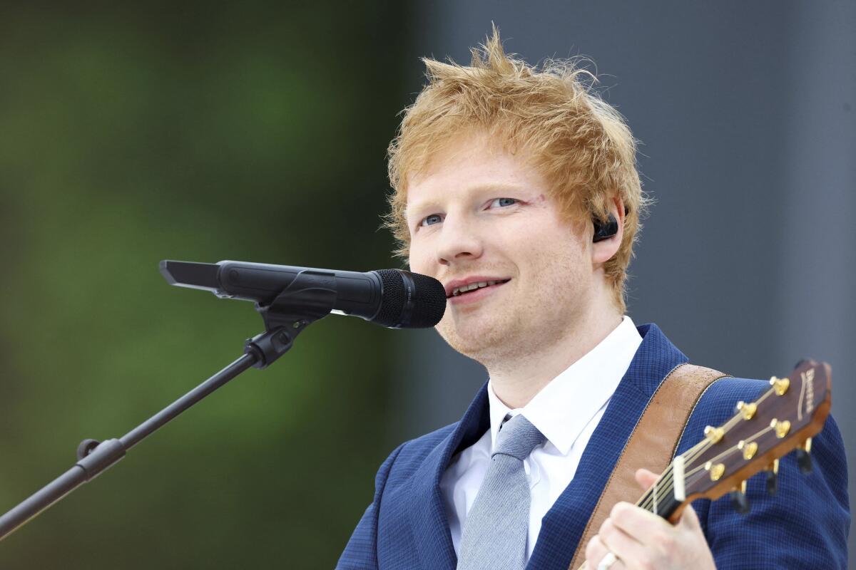 A red-haired man in a blue suit plays an acoustic guitar and sings into a microphone