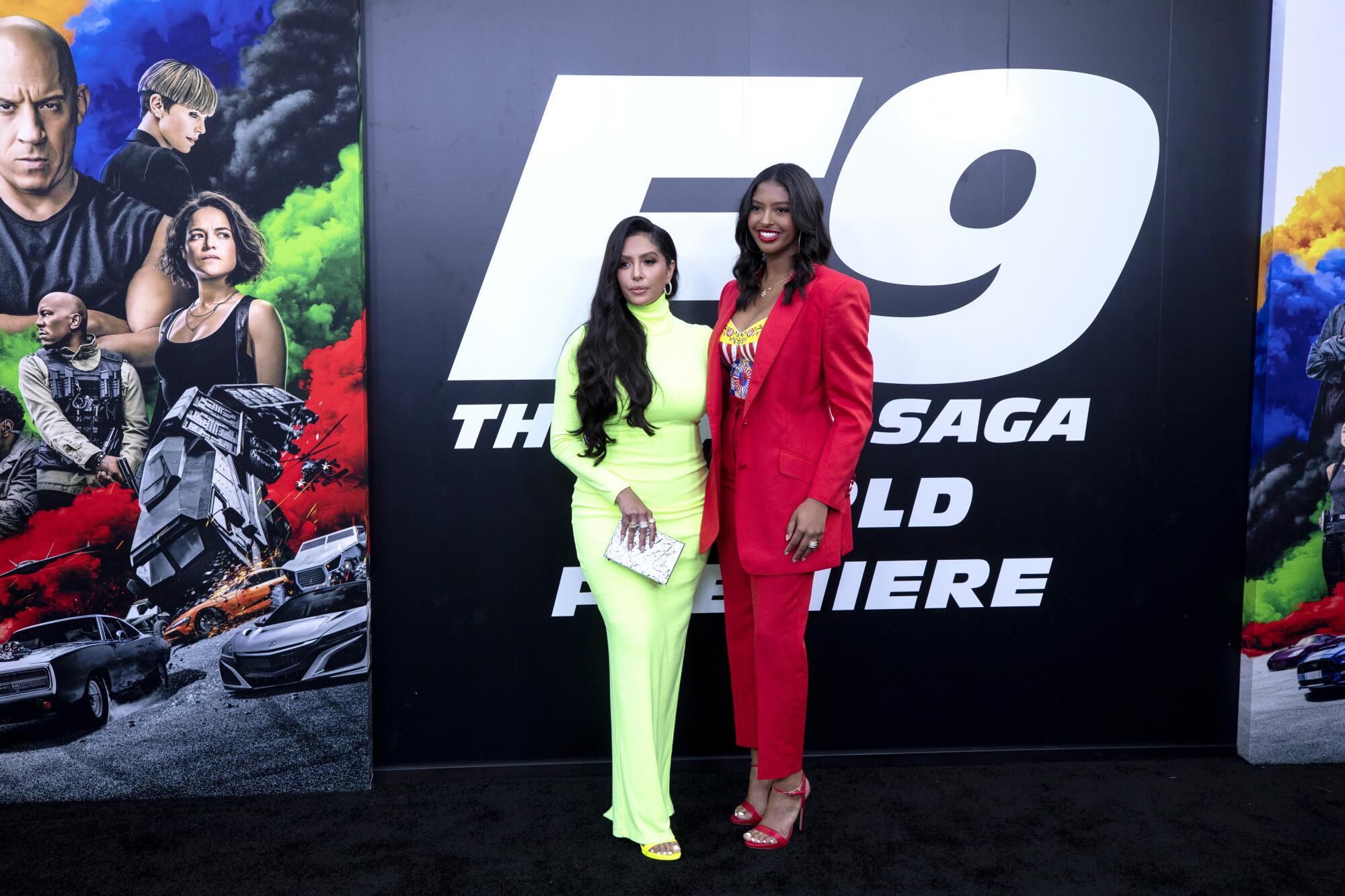 Vanessa Bryant in a bright yellow-green gown and daughter Natalia in a red suit stand in front of the "F9" premiere sign