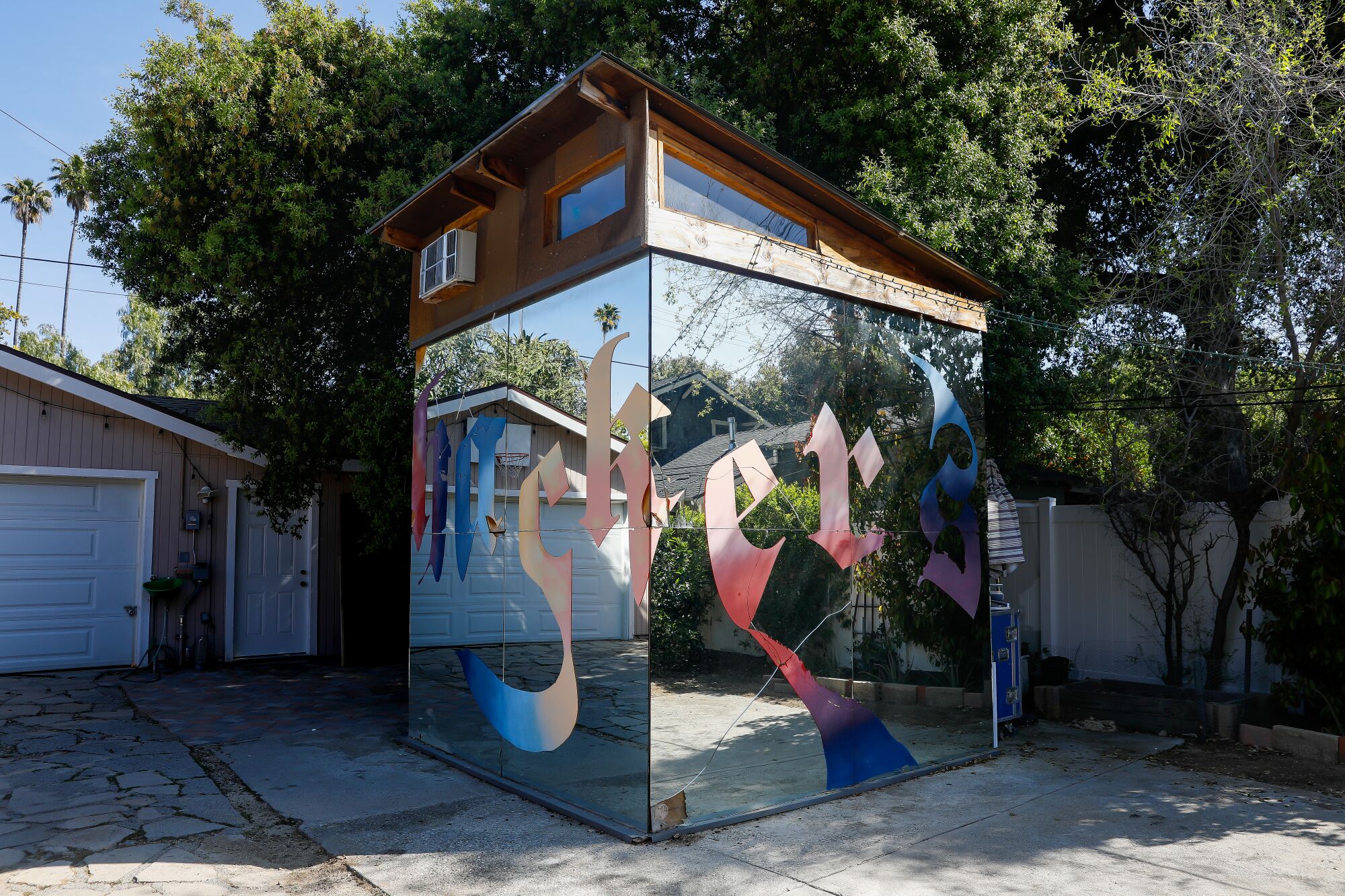 A view of a mirrored cube in a backyard in Pasadena. The facade has multicolored letters in Old English script.
