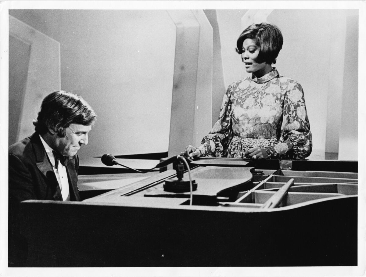 A seated Burt Bacharach plays the piano as Dionne Warwick sings.