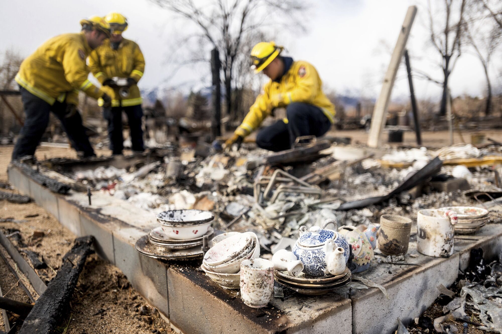 Small bowls, plates and a tea pot sit on the edge of the burned ruins of a home as firefighters look at the wreckage