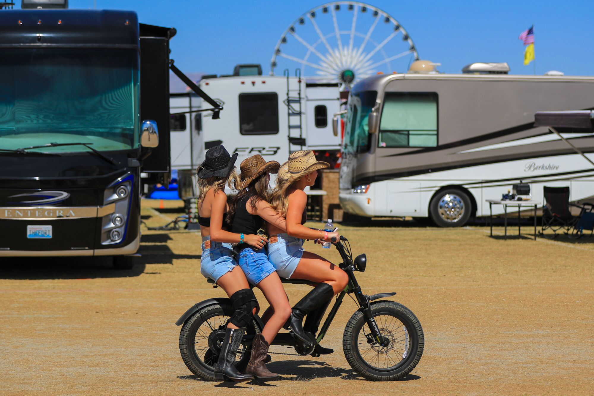 Three women in cowboy boots, hats and shorts ride electric bicycles in the Stagecoach campsite.