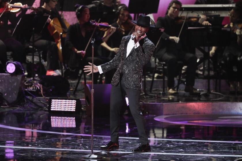 A man in a fancy suit and black hat sings into a microphone in front of seated orchestra-style musicians