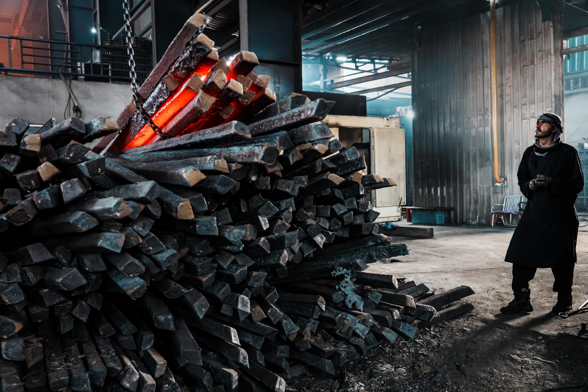 A man, right, looks up while standing near a big stack of metal rods in a warehouse-like setting