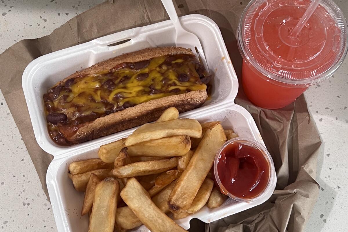 An open styrofoam container holds a vegan link with vegan chili and cheese and steak fries, with red Playa Punch on the side