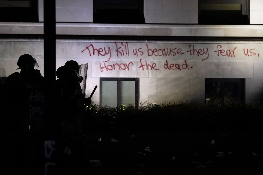 Police in riot gear stand in a line against protesters next to a message spay painted on the Kenosha County Courthouse, late Monday, Aug. 24, 2020, in Kenosha, Wis. Protesters converged on the county courthouse during a second night of clashes after the police shooting of Jacob Blake a day earlier turned Kenosha into the nation's latest flashpoint city in a summer of racial unrest. (AP Photo/David Goldman)