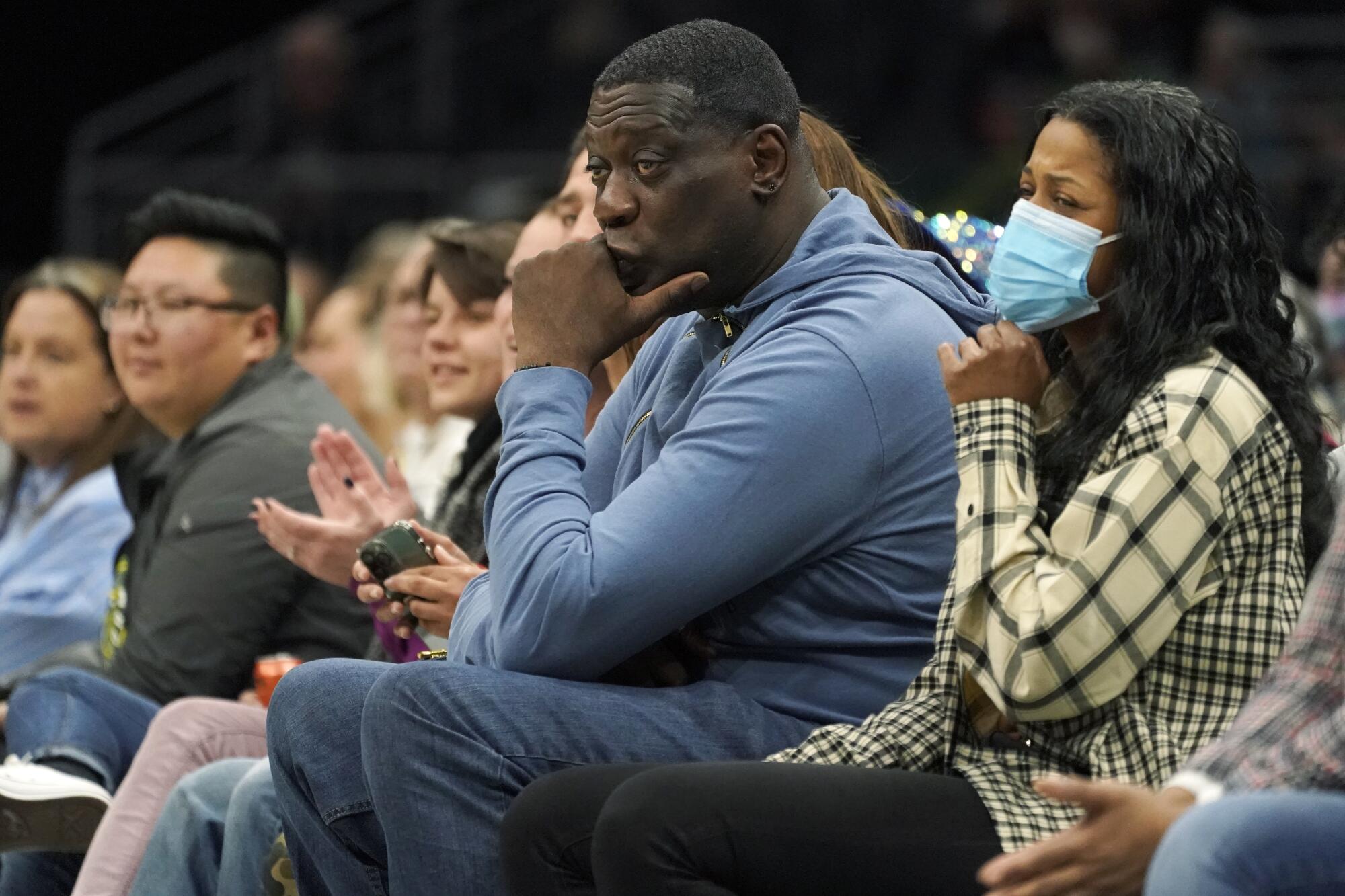 Shawn Kemp, wearing all blue, sits in courtside seats with his chin in his hand.