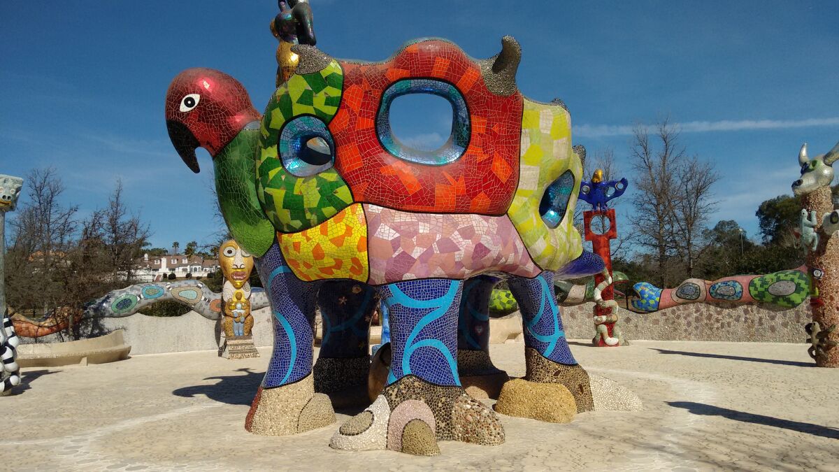 A multicolored sculpture covered in mosaic patterns stands outdoors among similar art pieces.